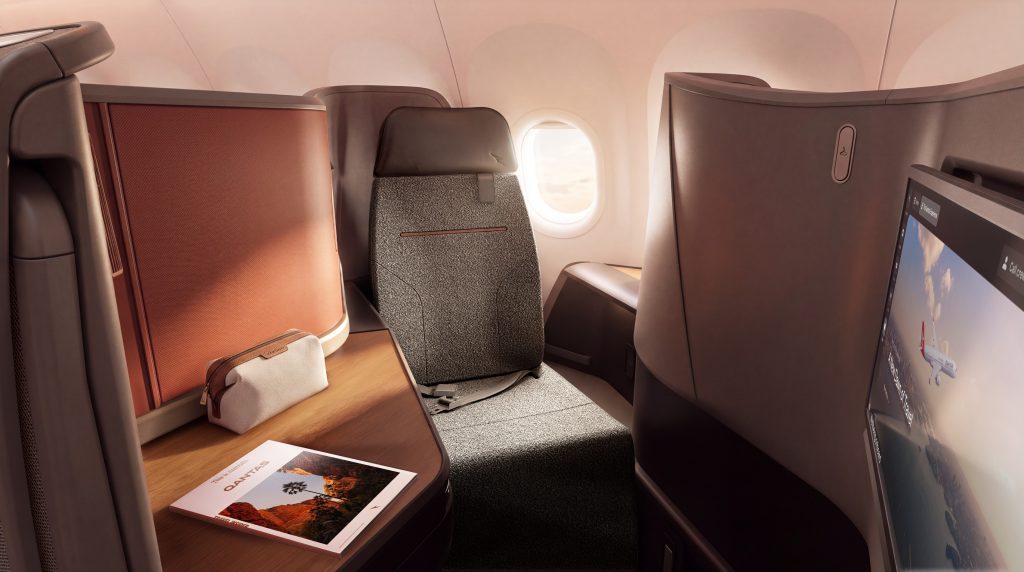 Business-class airplane cabin interior with a private seat, window view, and a large personal screen on the right; a brochure lies on the wooden table.