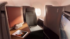 Business-class airplane cabin interior with a private seat, window view, and a large personal screen on the right; a brochure lies on the wooden table.