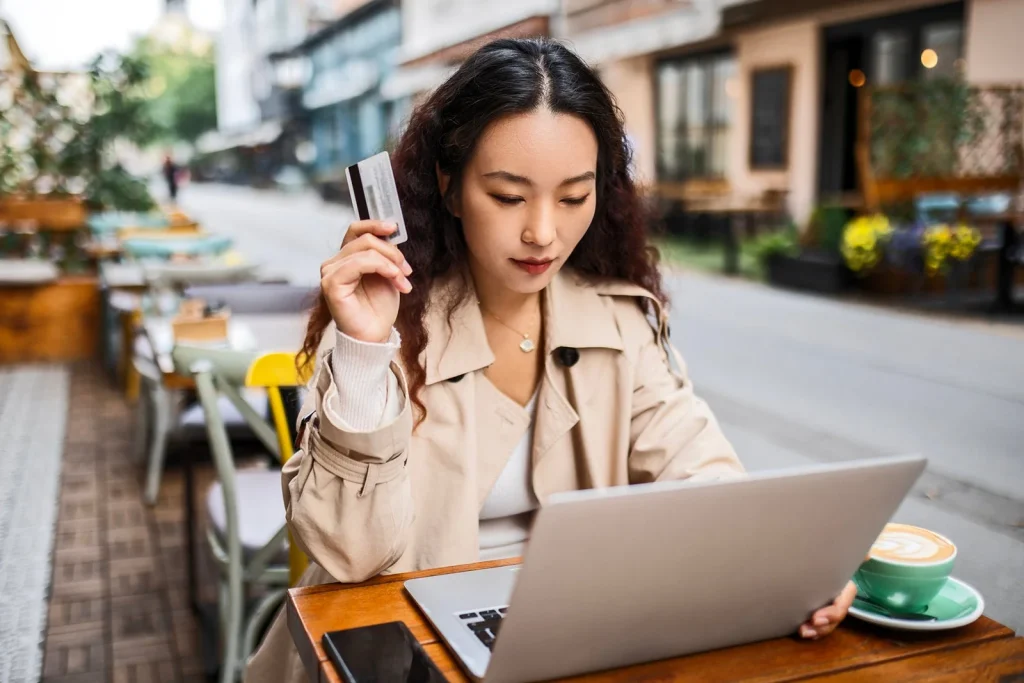 Woman at an outdoor cafe using a laptop, holding a credit card near her face as she looks at the screen.