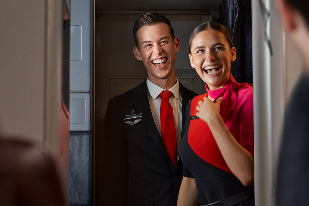 Two smiling flight attendants stand in an airplane cabin doorway, wearing professional uniforms with red accents, greeting passengers enthusiastically, warm teamwork vibe.