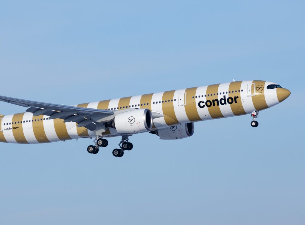 Condor passenger jet with gold-and-white striped livery and landing gear down in flight against a blue sky