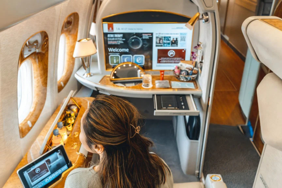 Private jet cabin interior with a passenger at a wood desk, using a tablet while a large screen shows a welcome page.